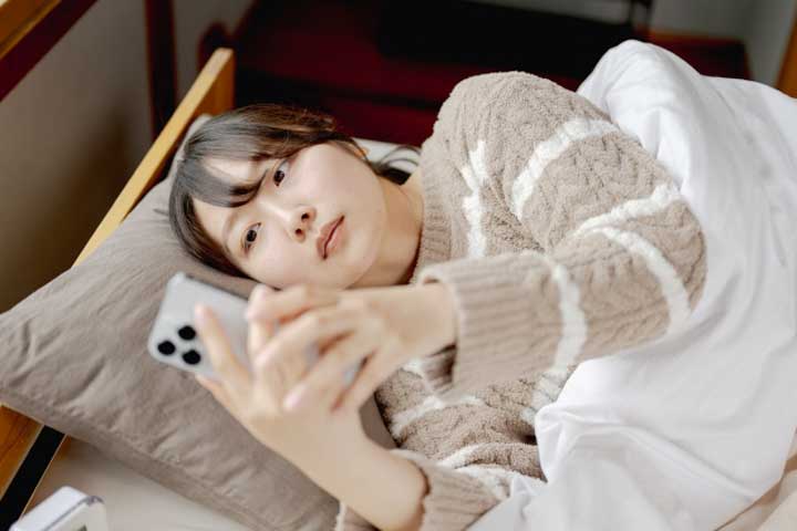 寝床でスマホをしながら横になる女性